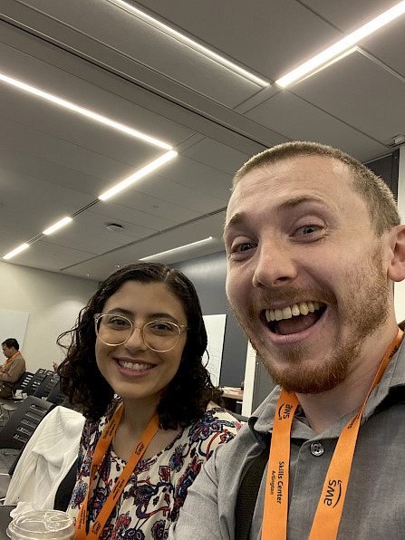Joe is on the right, Lili is on the left, both sitting and smiling in a conference room with orange lanyards that say Amazon.