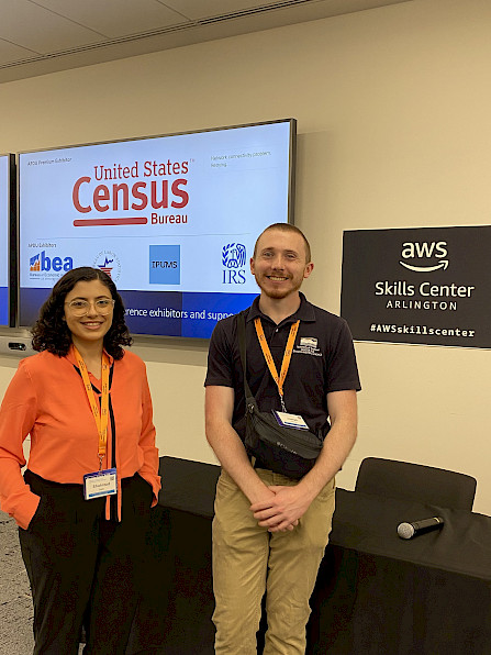 Lili on the left, Joe on the right, standing in front of a screen that lists U.S. agencies sponsoring the conference, like the U.S. Census, and a wall frame that says Amazon Skills Center.