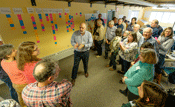 Participants stand around Bill Dennison who has his back to a wall. The wall has values of the watershed (such as history, beauty, and water supply) written on white index cards. Below each index card are multi-colored post it notes. People are smiling an