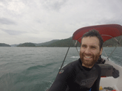 Jason is shown sitting on a boat in front of forested islands and mountains while he was traveling to a research site in Ilha Grande, Brazil.