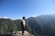 Angie Wei stands on a rocky outcrop overlooking a forested mountain ridge behind her.