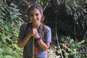 Yesenia appears smiling in the middle of the image surrounded by lush green forest, including low-growing shrubs. You can see her upper body and her hands are holding a walking stick in front of her body.
