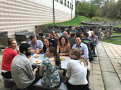25 people sit on a patio, spread out over 3 picnic tables. The patio is next to a large, brick building and a grassy area with low, rock walls.