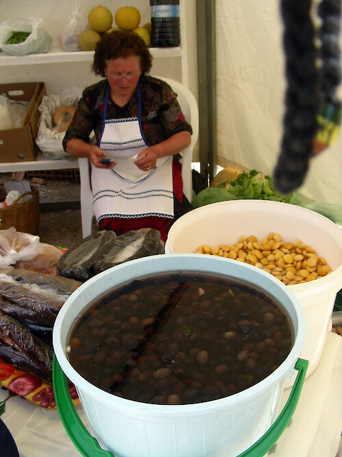 A hillside village in the upland Algarve region of southwestern Portugal hosts a country fair with agricultural and farm products for sale. Here a woman is selling olives and fava beans, as well as sausages.