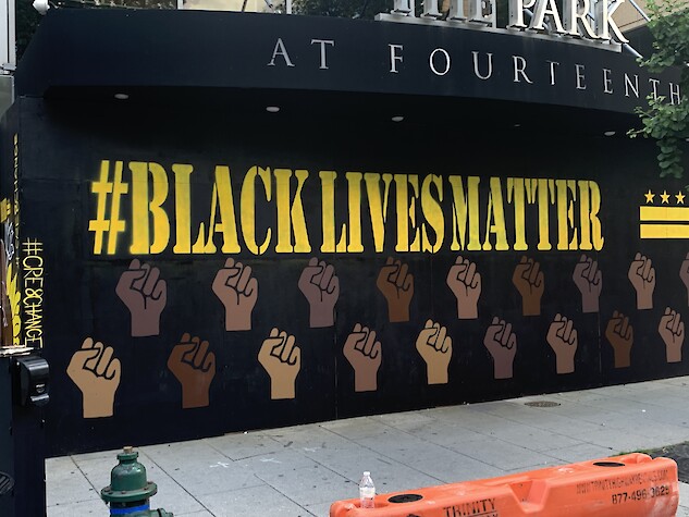 A painted mural that says #BlackLivesMatter in yellow wording. The background of the mural is black. The Washington D.C. flag is painted in yellow on the right- hand corner. Below the #BlackLivesMatter writing there are Black power fists in various shades of brown skin tones.