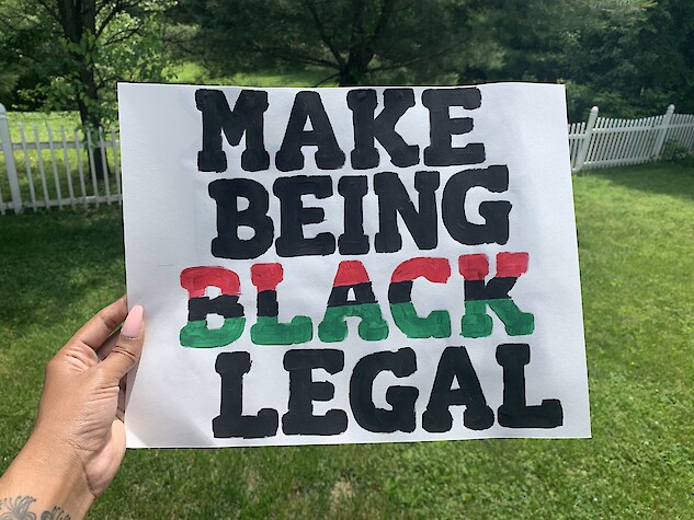 A handmade protest sign that says “Make Being Black Legal.” The word Black is painted using the colors of the African-American flag (red, black, and green). The remaining letters are painted black.