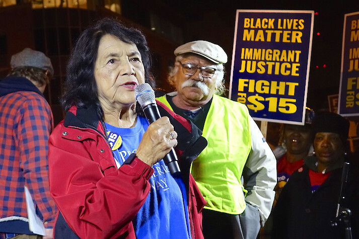 Dolores Huerta speaks over a microphone at an event; a man is next to her and others can be seen standing behind her. A sign in the background reads “Black Lives Matter, Immigrant Justice, Fight for $15.”
