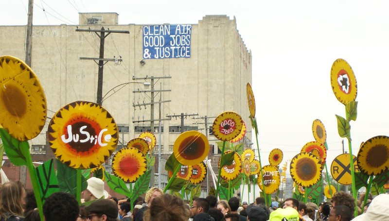 Sunflower signs are held up at a rally in Detroit and a banner reading “clean air, good jobs, and justice” is displayed on a building.