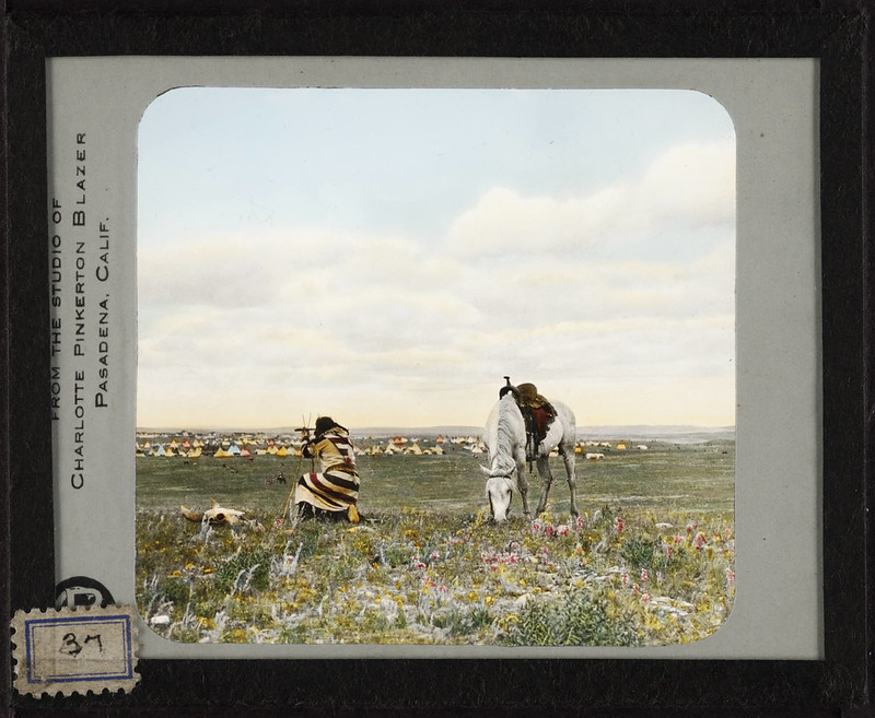 A native man in traditional native clothing kneeling on the ground looking through a spy glass supported by a twig made tripod. There is a cattle skull a few feet from him. He is looking upon a varying and brightly colored teepee camp, and his white horse, fully tacked, is  standing behind him grazing.