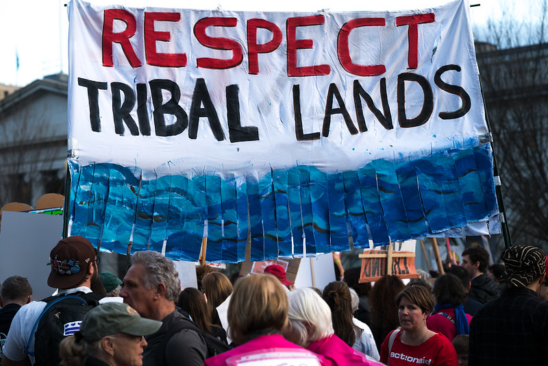 A cloth protest sign with the words “Respect Tribal Lands” painted in red and all capital letters. The sign is supported by two wood poles, and the bottom of the sign is painted  with water waves of varying blues and fringed.