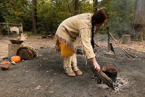 A woman with dark hair and skin dressed in traditional native clothing and facial paint, bending over a pot that’s on a smoldering fire. She is holding a log.