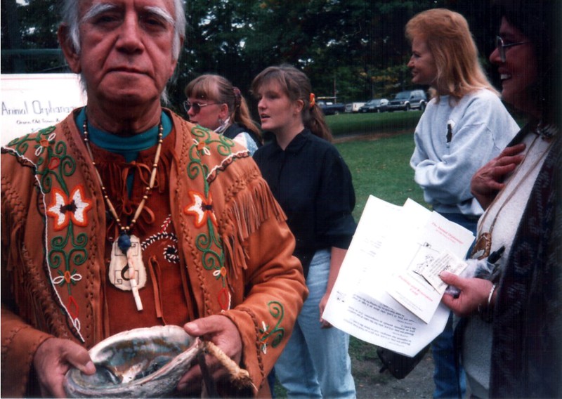 A man with medium-colored skin and white hair wearing a traditional animal skin shirt with fringe. He is holding an large iridescent shell with the concave side up. There are four other people in the background smiling with their attention drawn somewhere else.
