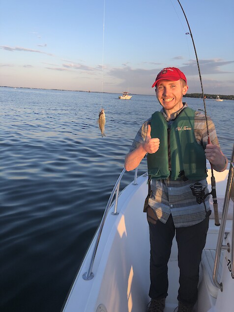 Joe Edgerton is standing on one side of the fishing boat and is holding his fishing pole with a small, five or so inch perch fish on the end of the fishing line.