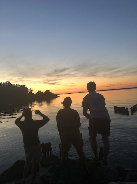 Dark silhouettes of three people are seen standing on some rocks while one of them is fishing in open water during a sunset.
