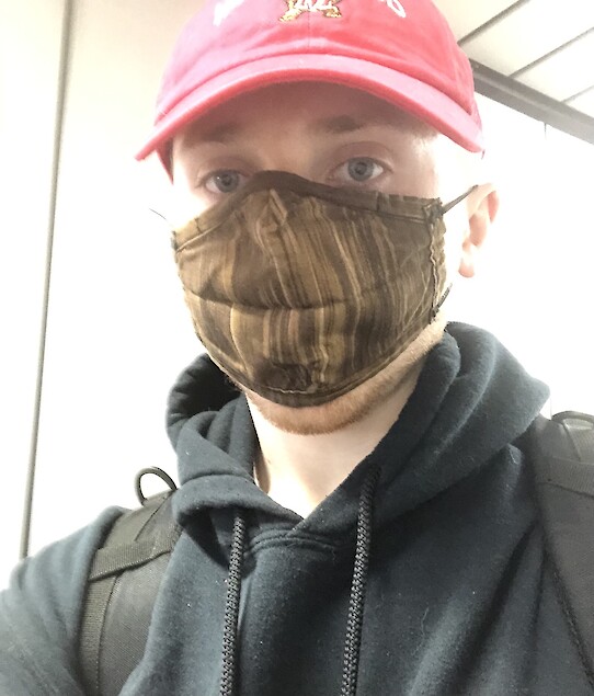 Selfie of Joseph Edgerton wearing a black sweatshirt along with a red University of Maryland baseball hat as he is walking off of a plane and into an airport terminal.