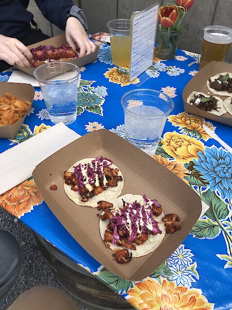 The legs of three people sitting around a table with different dishes and drinks in front, including a plate of cauliflower tacos and a side of potato chips, both covered in Old Bay Seasoning.