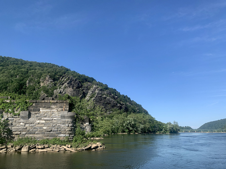 The Potomac and Shenandoah Rivers converge at Harper’s Ferry National Historic Park. To the side of the calm converging rivers, there is a large foothill and rock face, in addition to stone barriers placed in the center of the Potomac side of the tributary.