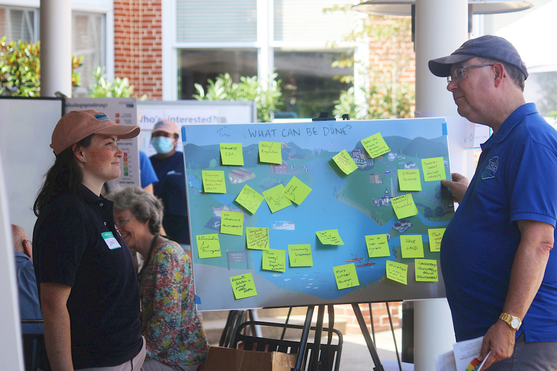 Sidney and a participant are facing each other and engaged in conversation with a conceptual diagram poster standing between them. The poster board reads at the top “What can be done?” and is filled with green sticky notes of responses.