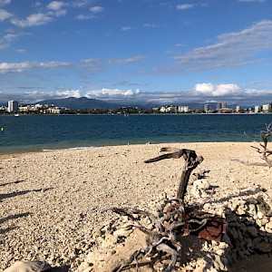 A pale beach of corals in the foreground; in the background, a blue lagoon and a city of pale buildings that hugs the coastline. There is a mountain in the far background.