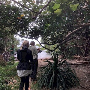 Two European woomen walk through a mangrove forest. The branches hand low overhead. The ocean is just visible in the background.
