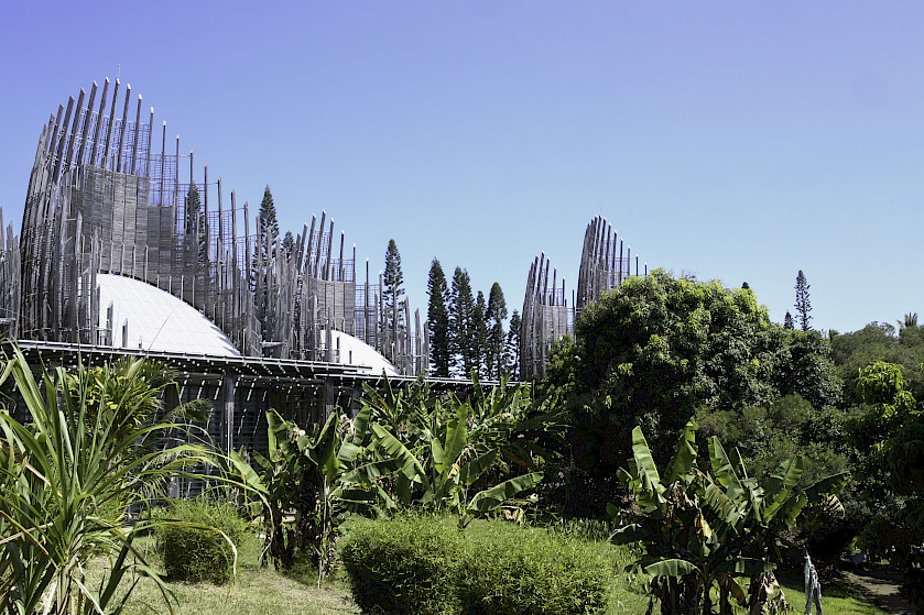 From a tropical jungle treeline, silvery structures resembling woven huts rise into a pale blue sky.