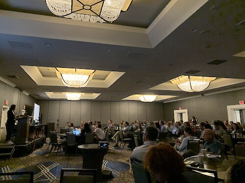 Photo of a large audience seated at tables in a spacious room during the 'Beyond 2025: Visionary Paths in the Chesapeake Bay Restoration by the Next Generation' panel discussion at the Chesapeake Community Research Symposium. The attendees are focused on the speakers, demonstrating engagement with the topic.