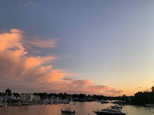 Photo of a sunset viewed from the Spa Creek Drawbridge in Annapolis, MD, featuring boats in both the foreground and background. The image captures the colorful sky at dusk, with the silhouettes of boats adding to the scenic waterway ambiance.