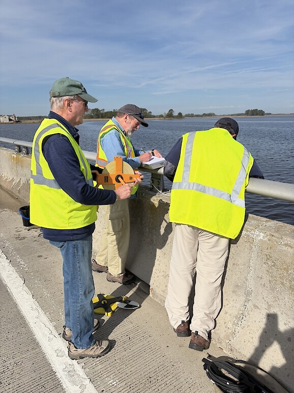 Three men in yellow vests stand beside the edge of a bridge and engage in water monitoring.